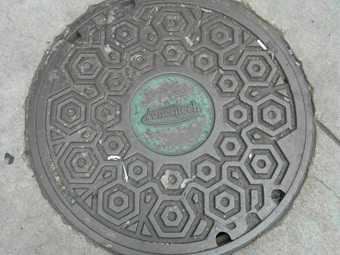 A round manhole cover on a concrete surface featuring a pattern of hexagons. The center of the cover is marked with the word 'Ameritech' in raised letters, surrounded by a slightly greenish patina. The cover has a few visible scratches and wear marks, suggesting it has been in use for some time.