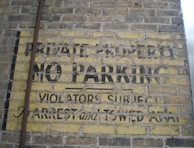 An old and weathered sign painted on a brick wall with the words 'PRIVATE PROPERTY NO PARKING VIOLATORS SUBJECT TO ARREST and TOWED AWAY'. The paint is faded and peeling, revealing the yellow background with black text.