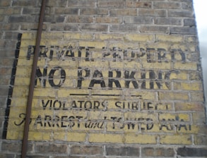An old and weathered sign painted on a brick wall with the words 'PRIVATE PROPERTY NO PARKING VIOLATORS SUBJECT TO ARREST and TOWED AWAY'. The paint is faded and peeling, revealing the yellow background with black text.