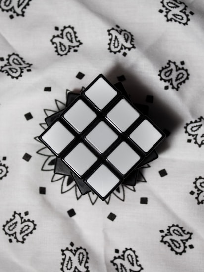 A solved Rubik's Cube with all sides showing white squares is placed on a patterned white fabric background. The cube appears to be in a classic 3x3 configuration, and the fabric features a repeating paisley and geometric design in black.