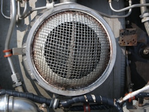 A metallic circular grill is surrounded by industrial pipes and fittings. The scene includes metal tubes, some wrapped with black insulation and others with metallic and orange bands. The environment appears to be part of a mechanical or industrial setup, featuring various bolts and a rusted rectangular plate.