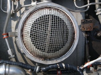 A metallic circular grill is surrounded by industrial pipes and fittings. The scene includes metal tubes, some wrapped with black insulation and others with metallic and orange bands. The environment appears to be part of a mechanical or industrial setup, featuring various bolts and a rusted rectangular plate.