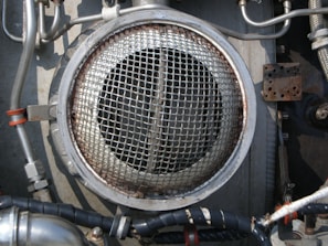 A metallic circular grill is surrounded by industrial pipes and fittings. The scene includes metal tubes, some wrapped with black insulation and others with metallic and orange bands. The environment appears to be part of a mechanical or industrial setup, featuring various bolts and a rusted rectangular plate.
