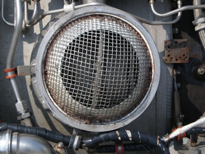 A metallic circular grill is surrounded by industrial pipes and fittings. The scene includes metal tubes, some wrapped with black insulation and others with metallic and orange bands. The environment appears to be part of a mechanical or industrial setup, featuring various bolts and a rusted rectangular plate.