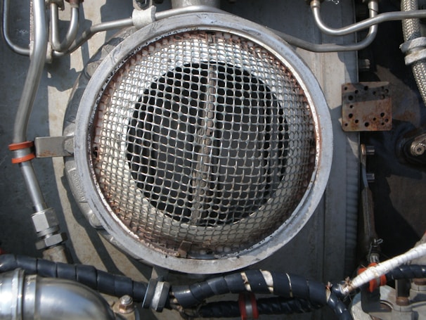 A metallic circular grill is surrounded by industrial pipes and fittings. The scene includes metal tubes, some wrapped with black insulation and others with metallic and orange bands. The environment appears to be part of a mechanical or industrial setup, featuring various bolts and a rusted rectangular plate.