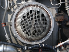 A metallic circular grill is surrounded by industrial pipes and fittings. The scene includes metal tubes, some wrapped with black insulation and others with metallic and orange bands. The environment appears to be part of a mechanical or industrial setup, featuring various bolts and a rusted rectangular plate.