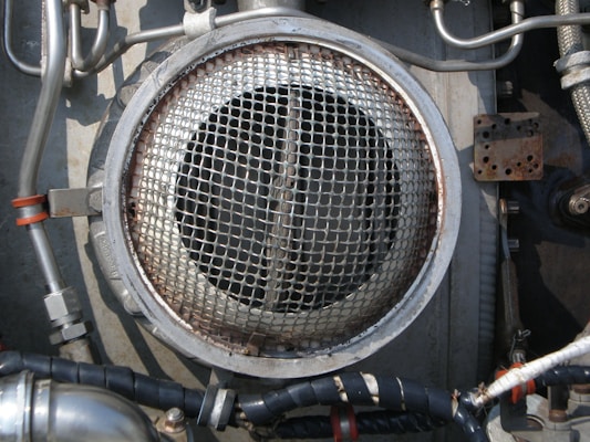 A metallic circular grill is surrounded by industrial pipes and fittings. The scene includes metal tubes, some wrapped with black insulation and others with metallic and orange bands. The environment appears to be part of a mechanical or industrial setup, featuring various bolts and a rusted rectangular plate.