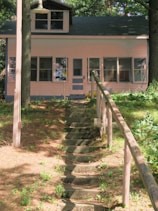 A small, charming house surrounded by trees and greenery. A stone pathway with rustic wooden railings leads up a gentle slope to the front door. The house features large windows and is painted in a soft, light color.