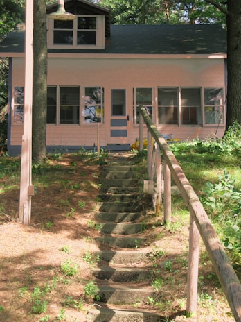 A small, charming house surrounded by trees and greenery. A stone pathway with rustic wooden railings leads up a gentle slope to the front door. The house features large windows and is painted in a soft, light color.