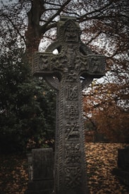 An intricately carved stone cross stands in a graveyard setting, surrounded by fallen autumn leaves and dense foliage. The cross features detailed Celtic designs and symbols, with a backdrop of bare tree branches.
