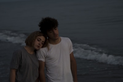A young couple stands on a beach during an overcast day. The woman rests her head on the man's shoulder, conveying a sense of closeness and calm. Gentle waves can be seen in the background under a dim sky.