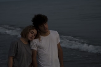 A young couple stands on a beach during an overcast day. The woman rests her head on the man's shoulder, conveying a sense of closeness and calm. Gentle waves can be seen in the background under a dim sky.