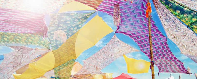 A canopy of vibrantly colored fabric panels stretches across the sky, featuring a variety of patterns including zigzags, florals, and abstract designs. Sunlight filters through the fabric, creating a bright and festive atmosphere.