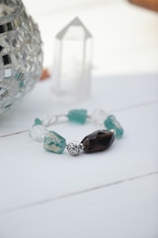 An intricately designed bracelet featuring a variety of crystal beads in blue and clear colors is placed on a white surface. Beside it is a decorative silver mosaic glass container and a clear, faceted quartz crystal tower.