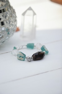 An intricately designed bracelet featuring a variety of crystal beads in blue and clear colors is placed on a white surface. Beside it is a decorative silver mosaic glass container and a clear, faceted quartz crystal tower.