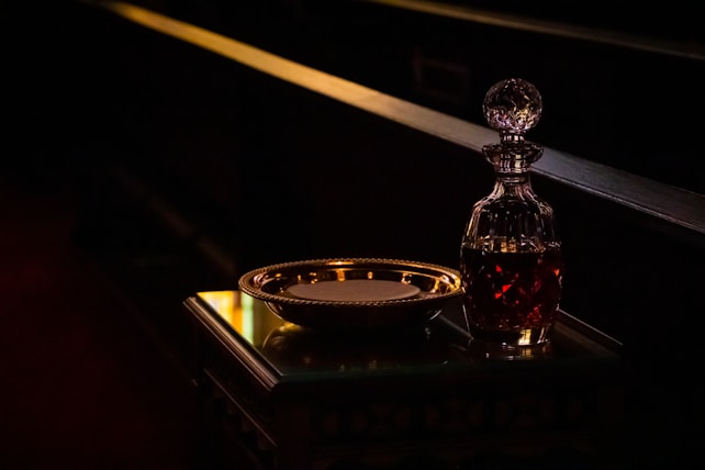 A crystal decanter filled with a dark liquid is placed on a polished wooden table, beside a gleaming gold-rimmed plate. The scene is dimly lit, casting reflections and shadows that create a warm, intimate atmosphere.