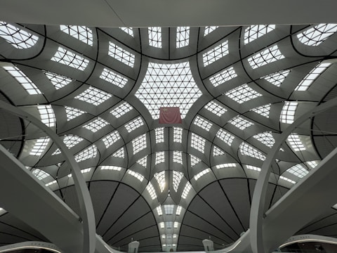 A modern architectural ceiling with an intricate design featuring curved and overlapping segments. The ceiling is made of metal and glass panels arranged in geometric patterns, allowing natural light to filter through. The structure is grand and spacious, with large beams supporting the design.