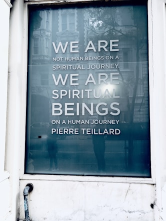 A large window displays a reflective surface with an inspiring quote reading 'WE ARE NOT HUMAN BEINGS ON A SPIRITUAL JOURNEY. WE ARE SPIRITUAL BEINGS ON A HUMAN JOURNEY.' The text is attributed to Pierre Teilhard. The surrounding scene includes reflections of a building facade and some indistinct figures, suggesting a city environment.