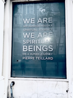 A large window displays a reflective surface with an inspiring quote reading 'WE ARE NOT HUMAN BEINGS ON A SPIRITUAL JOURNEY. WE ARE SPIRITUAL BEINGS ON A HUMAN JOURNEY.' The text is attributed to Pierre Teilhard. The surrounding scene includes reflections of a building facade and some indistinct figures, suggesting a city environment.