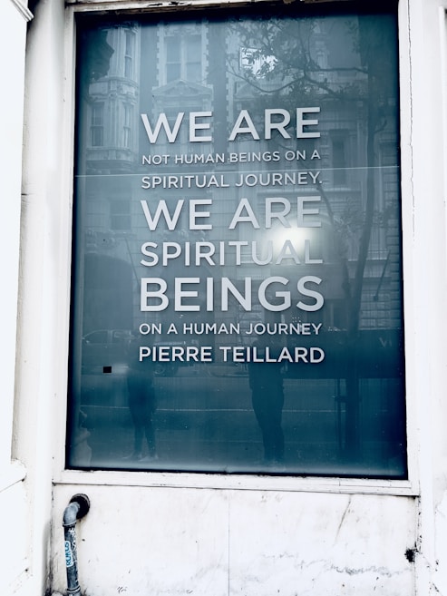 A large window displays a reflective surface with an inspiring quote reading 'WE ARE NOT HUMAN BEINGS ON A SPIRITUAL JOURNEY. WE ARE SPIRITUAL BEINGS ON A HUMAN JOURNEY.' The text is attributed to Pierre Teilhard. The surrounding scene includes reflections of a building facade and some indistinct figures, suggesting a city environment.