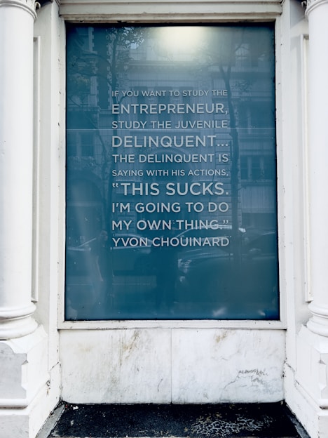 A glass window displays a quote about entrepreneurship and delinquency attributed to Yvon Chouinard. The text is written in a modern, sans-serif font on a teal-blue background. The window frame is white, and there are reflections visible on the glass.