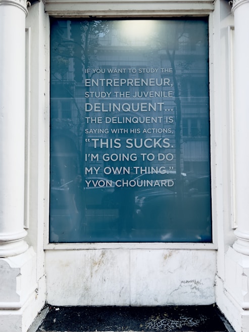 A glass window displays a quote about entrepreneurship and delinquency attributed to Yvon Chouinard. The text is written in a modern, sans-serif font on a teal-blue background. The window frame is white, and there are reflections visible on the glass.