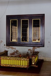 A traditional wooden musical instrument with ornate gold detailing and a row of yellow resonators is placed against a wall with visible signs of wear and peeling paint. Above it, a wooden framed window with vertical bars adds to the vintage aesthetic.
