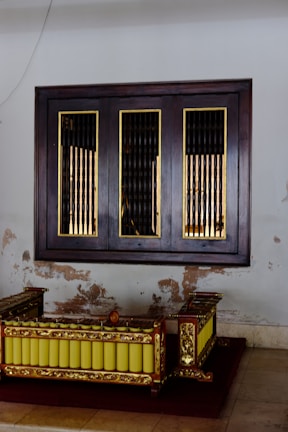 A traditional wooden musical instrument with ornate gold detailing and a row of yellow resonators is placed against a wall with visible signs of wear and peeling paint. Above it, a wooden framed window with vertical bars adds to the vintage aesthetic.