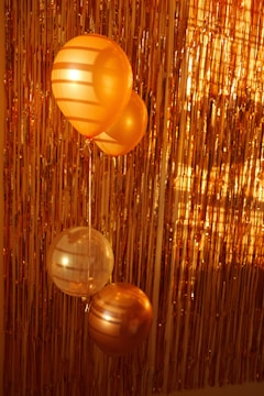 Three balloons, primarily in shades of gold and orange, are positioned against a backdrop of metallic tinsel. The lighting casts shadows on the balloons, enhancing their shiny and festive appearance.