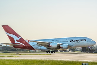 A large passenger aircraft with the Qantas logo is positioned on a runway, preparing for takeoff or having just landed. The airplane is predominantly white with a red tail featuring a white kangaroo logo. The background includes a clear sky and distant airport infrastructure.