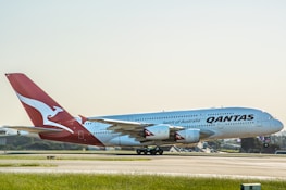 A large passenger aircraft with the Qantas logo is positioned on a runway, preparing for takeoff or having just landed. The airplane is predominantly white with a red tail featuring a white kangaroo logo. The background includes a clear sky and distant airport infrastructure.