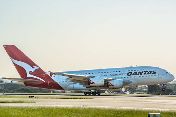 A large passenger aircraft with the Qantas logo is positioned on a runway, preparing for takeoff or having just landed. The airplane is predominantly white with a red tail featuring a white kangaroo logo. The background includes a clear sky and distant airport infrastructure.