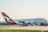 A large passenger aircraft with the Qantas logo is positioned on a runway, preparing for takeoff or having just landed. The airplane is predominantly white with a red tail featuring a white kangaroo logo. The background includes a clear sky and distant airport infrastructure.
