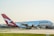 A large passenger aircraft with the Qantas logo is positioned on a runway, preparing for takeoff or having just landed. The airplane is predominantly white with a red tail featuring a white kangaroo logo. The background includes a clear sky and distant airport infrastructure.