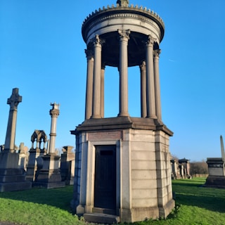 A historic cemetery with an ornate stone mausoleum featuring tall columns and a domed roof. Several other grave markers, including crosses and gothic stone structures, are present in the background. A clear blue sky enhances the serene atmosphere.