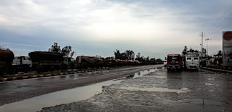 A wet, reflective road scene with two trucks parked on the side, under an overcast sky. There are several tanker trucks lined up along the opposite side of the road, bordered by greenery. A signboard on the right displays a logo associated with a petroleum company.