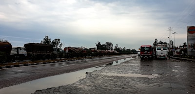 A wet, reflective road scene with two trucks parked on the side, under an overcast sky. There are several tanker trucks lined up along the opposite side of the road, bordered by greenery. A signboard on the right displays a logo associated with a petroleum company.