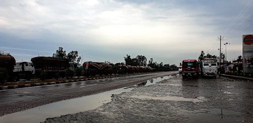A wet, reflective road scene with two trucks parked on the side, under an overcast sky. There are several tanker trucks lined up along the opposite side of the road, bordered by greenery. A signboard on the right displays a logo associated with a petroleum company.