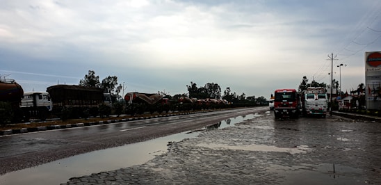 A wet, reflective road scene with two trucks parked on the side, under an overcast sky. There are several tanker trucks lined up along the opposite side of the road, bordered by greenery. A signboard on the right displays a logo associated with a petroleum company.