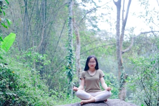 A person is meditating peacefully on a large rock amidst a lush forest. The surroundings are filled with dense green foliage, tall trees, and various types of plants, creating a serene natural setting.