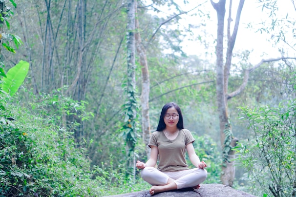 A person is meditating peacefully on a large rock amidst a lush forest. The surroundings are filled with dense green foliage, tall trees, and various types of plants, creating a serene natural setting.