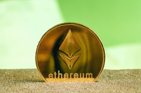 A gold-colored Ethereum coin is positioned upright on a textured surface with a blurred green background. The Ethereum logo and the word 'ethereum' are prominently displayed on the coin.