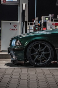 A dark green car is parked at a gas station next to a fuel pump. The focus is on the front side of the car, showing its headlight and wheel. The car is shiny and well-maintained, with sleek design details visible in the low lighting. The ORLEN logo is visible on the gas pump, which is positioned in the background under artificial lighting.