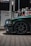A dark green car is parked at a gas station next to a fuel pump. The focus is on the front side of the car, showing its headlight and wheel. The car is shiny and well-maintained, with sleek design details visible in the low lighting. The ORLEN logo is visible on the gas pump, which is positioned in the background under artificial lighting.