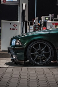 A dark green car is parked at a gas station next to a fuel pump. The focus is on the front side of the car, showing its headlight and wheel. The car is shiny and well-maintained, with sleek design details visible in the low lighting. The ORLEN logo is visible on the gas pump, which is positioned in the background under artificial lighting.
