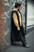 A person leans against a graffiti-covered gray wall, wearing a long black coat, beige hoodie, and dark pants. The scene features a mix of urban and casual style, with the person gazing to the side and wearing sunglasses.
