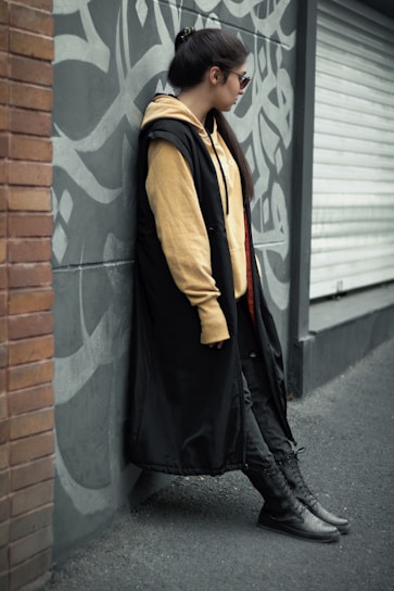 A person leans against a graffiti-covered gray wall, wearing a long black coat, beige hoodie, and dark pants. The scene features a mix of urban and casual style, with the person gazing to the side and wearing sunglasses.
