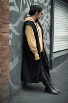 A person leans against a graffiti-covered gray wall, wearing a long black coat, beige hoodie, and dark pants. The scene features a mix of urban and casual style, with the person gazing to the side and wearing sunglasses.