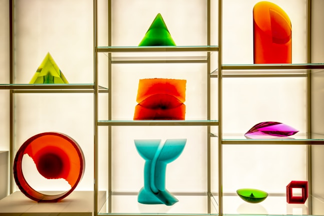 A display case features a variety of colorful geometric glass sculptures including pyramids, a circular ring, an abstract wave form, and other abstract shapes. The sculptures are composed of vibrant hues such as red, green, orange, blue, and purple and are placed on glass shelves against a softly lit background.