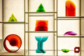 A display case features a variety of colorful geometric glass sculptures including pyramids, a circular ring, an abstract wave form, and other abstract shapes. The sculptures are composed of vibrant hues such as red, green, orange, blue, and purple and are placed on glass shelves against a softly lit background.